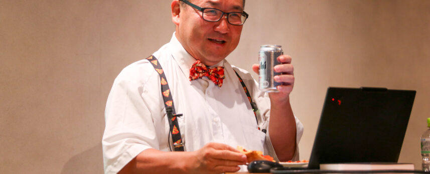 Man in a white shirt, red bow tie, and suspenders holds a can while eating pizza at a table with a laptop nearby, likely giving a presentation.