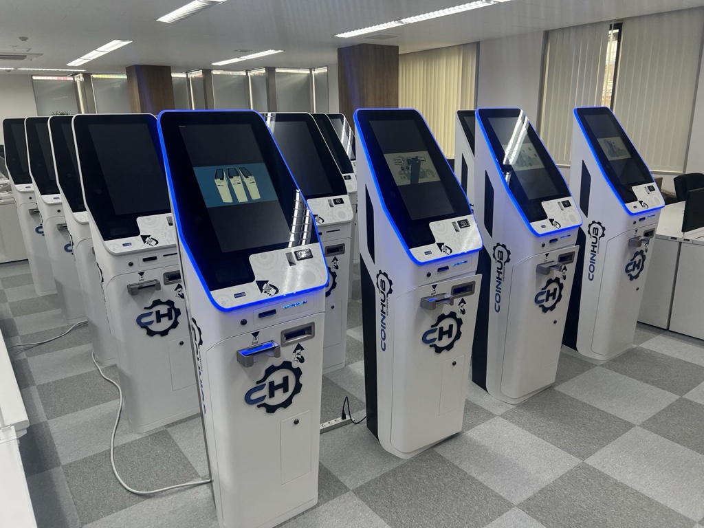 Row of white crypto ATM kiosks with blue LED edges in an office space, each with a touchscreen display.