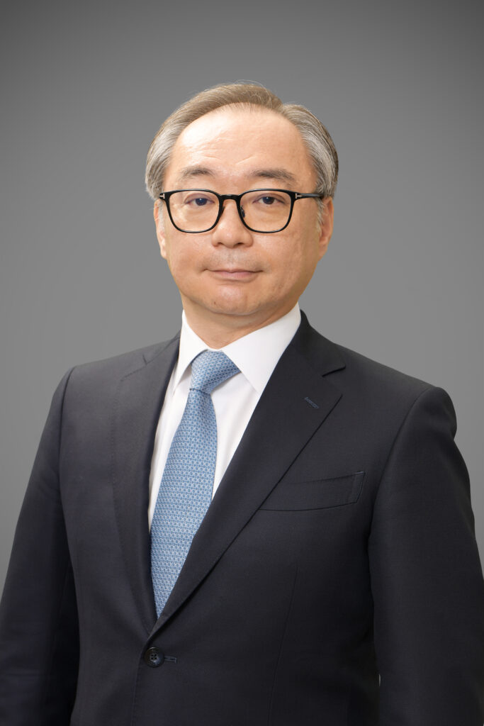 Professional head-and-shoulders portrait of a middle-aged Asian man in a dark suit, white shirt, and light blue tie, against a gray gradient background with a neutral expression.