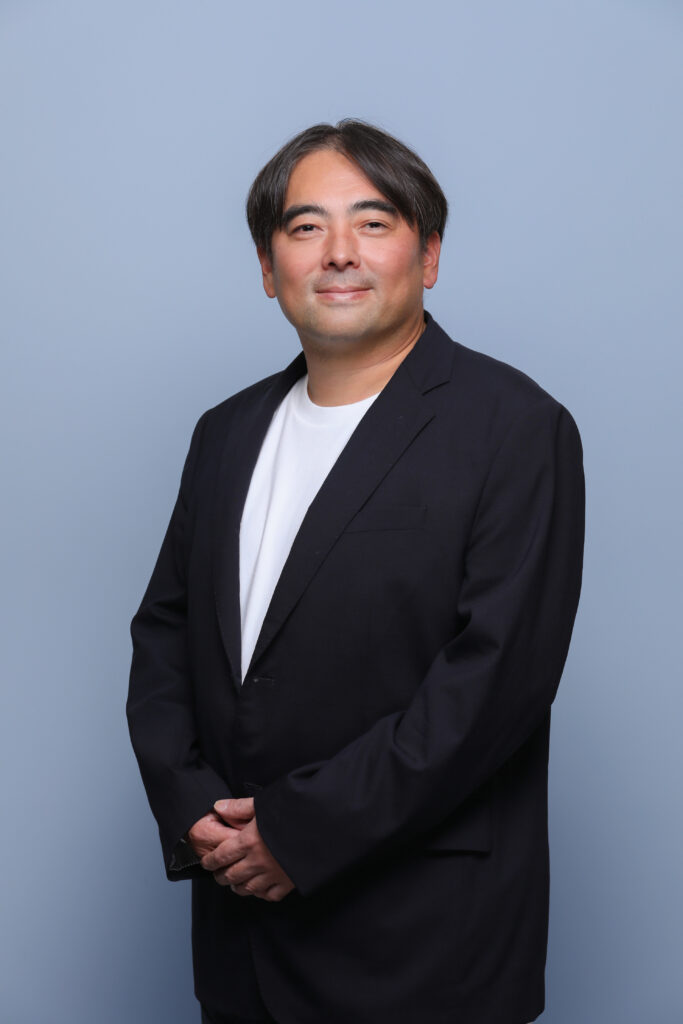 Professional headshot of a man with short dark hair, wearing a dark blazer and white shirt, standing against a blue background and smiling subtly.
