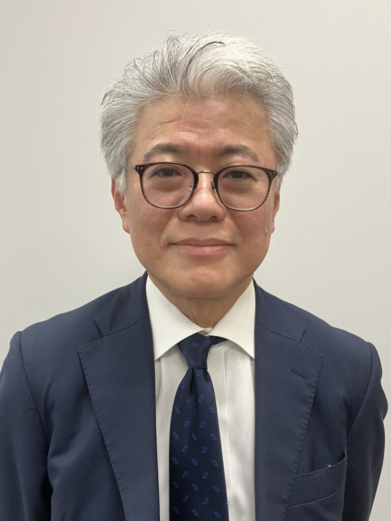 Close-up portrait of an older Asian man in a navy suit, white shirt, and blue patterned tie, wearing round glasses.