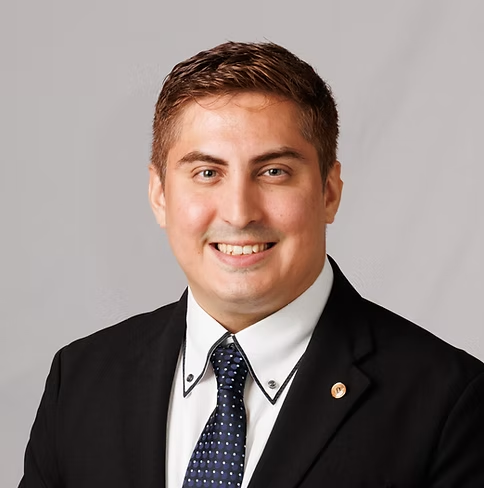Smiling man in a dark suit and blue tie, posing for a professional headshot against a gray background.