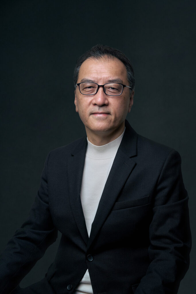Portrait of a middle-aged Asian man wearing glasses and a dark blazer against a dark studio backdrop.