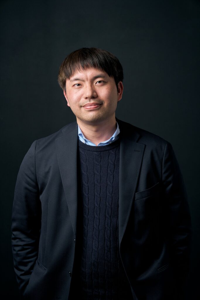 Portrait of an Asian man in a navy blazer and blue sweater, posing against a dark studio background with a gentle smile.