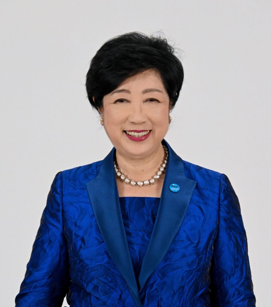 Smiling Asian woman in a bright blue jacket and pearl necklace, facing the camera against a plain light background.