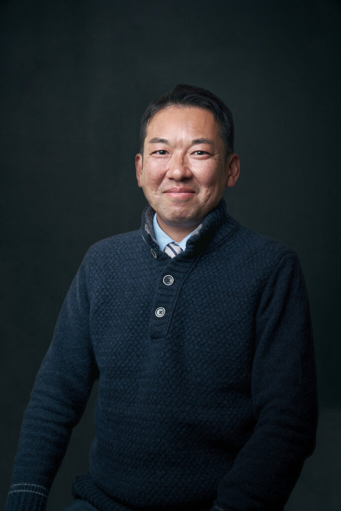 Portrait of a smiling man wearing a dark blue knit sweater over a light blue shirt and striped tie, posed against a dark background.
