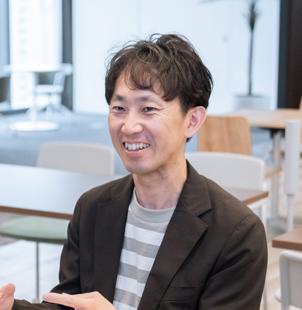 Smiling Asian man in a brown blazer and gray-striped shirt, seated at a table in a bright indoor space.